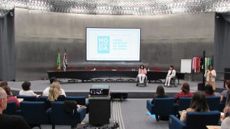 Imagem do auditório da Secretaria de Estado dos Direitos da Pessoa com Deficiência de São Paulo. Em primeiro plano, alunos sentados em poltronas. Ao fundo, no palco do auditório, a Secretária Célia Leão e a gerente do Programa Moda Inclusiva Izabelle Palma.
