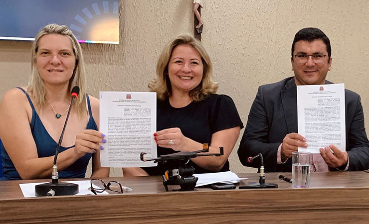 Fotografia colorida. Secretária Aracélia Costa e outras duas autoridades sentadas próximas a uma mesa segurando um documento. Ambas olham para a câmera com um sorriso no rosto.